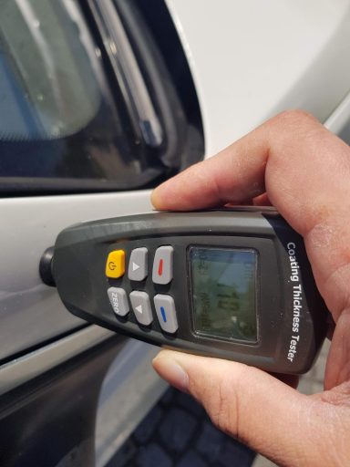 Hände halten ein Messgerät, das an einem Autolack getestet wird.